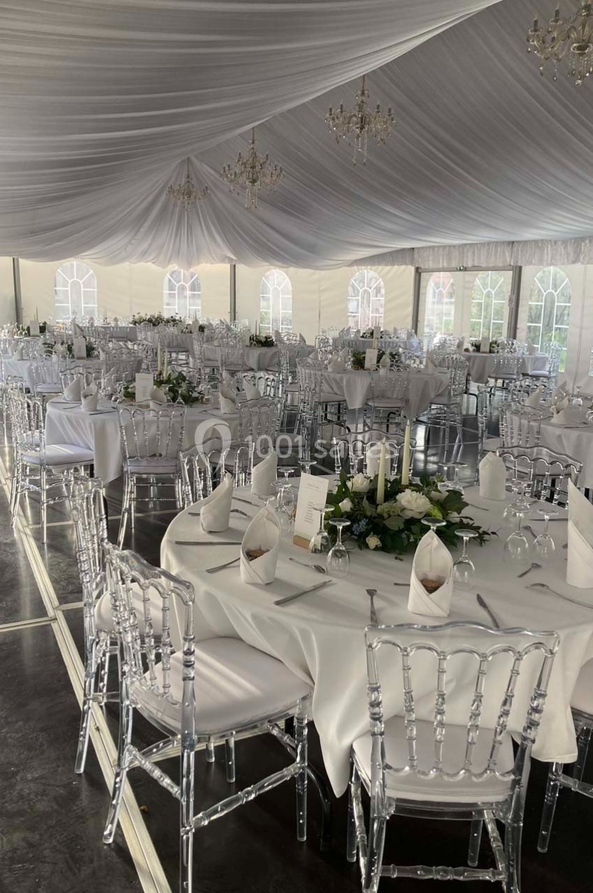 Salle de réception sous chapiteau avec tables rondes dressées, nappes blanches et décorations florales.