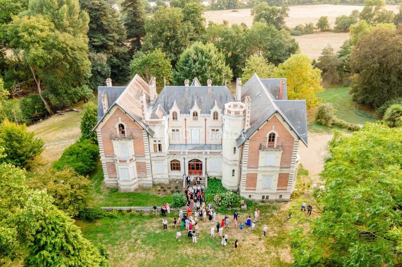 Vue aérienne d'un château ancien entouré de verdure, avec un groupe de personnes rassemblées devant l'entrée.