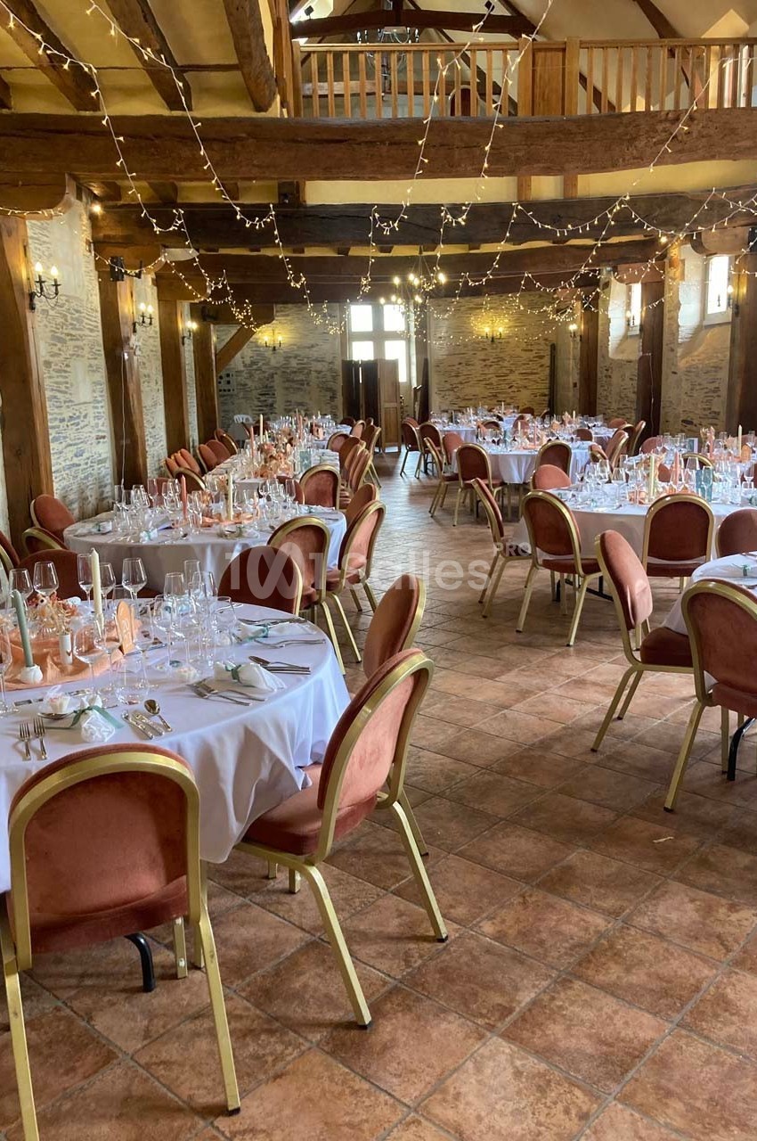 Salle de réception décorée avec des tables rondes dressées, chaises rouges et guirlandes lumineuses suspendues.
