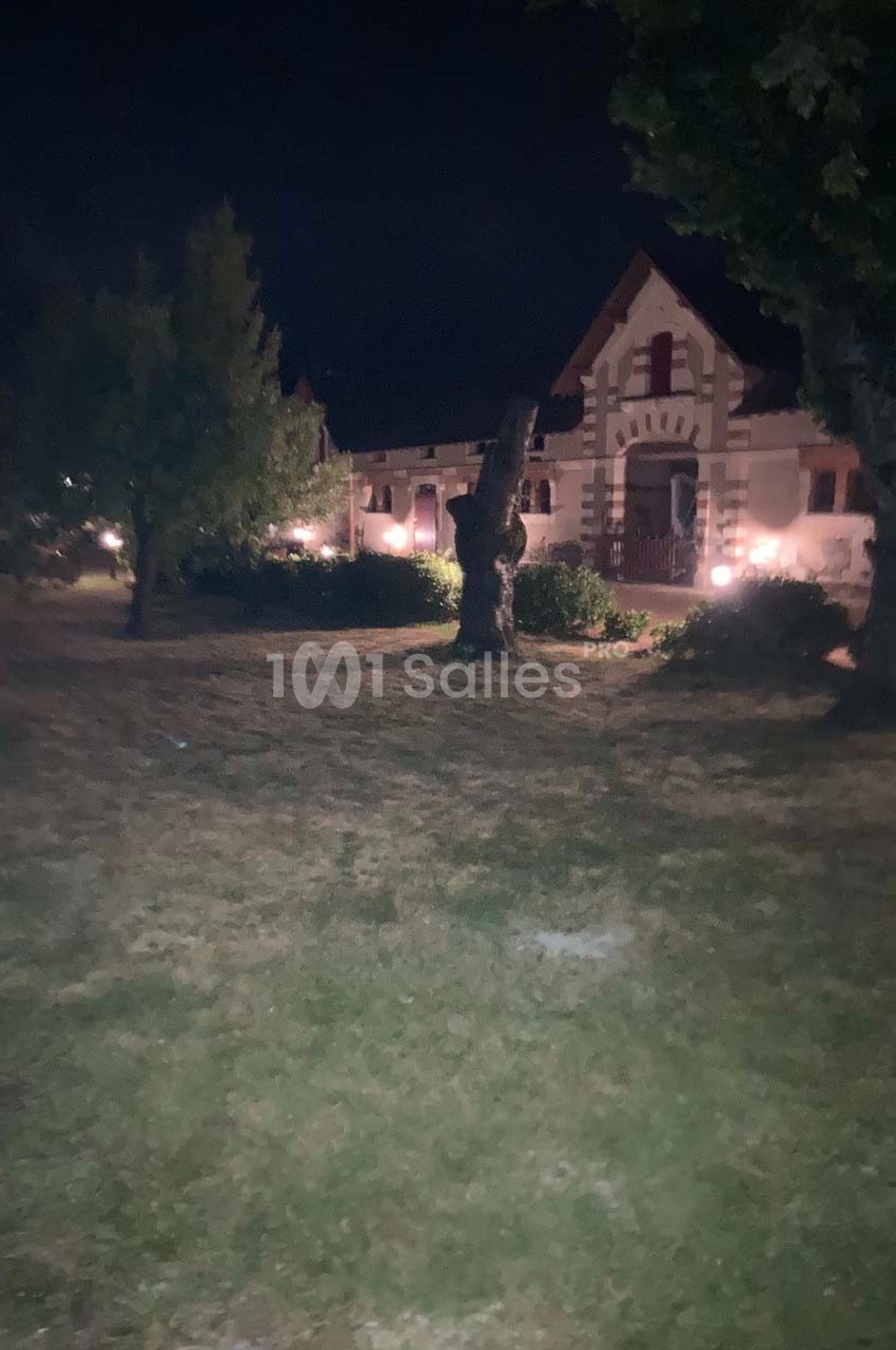Maison ancienne éclairée la nuit, entourée d'arbres et d'un jardin.