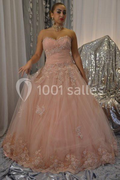 Femme portant une robe de soirée rose ornée de broderies, posant devant un fond argenté et des rideaux.