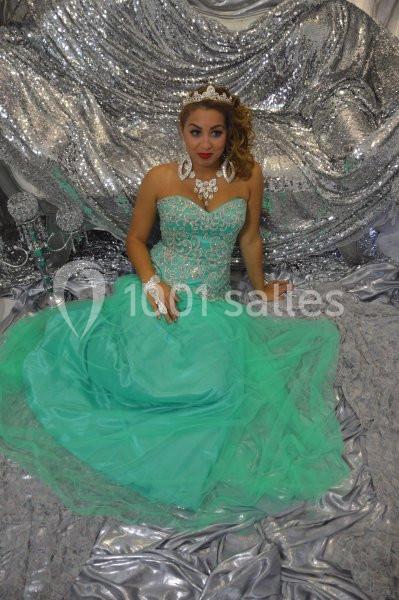 Femme en robe verte élégante, assise sur un fond argenté scintillant, portant des bijoux et une couronne.