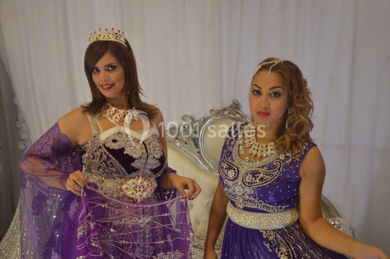 Deux femmes en robes traditionnelles violettes ornées de bijoux, posant devant un fond clair avec un fauteuil argenté.