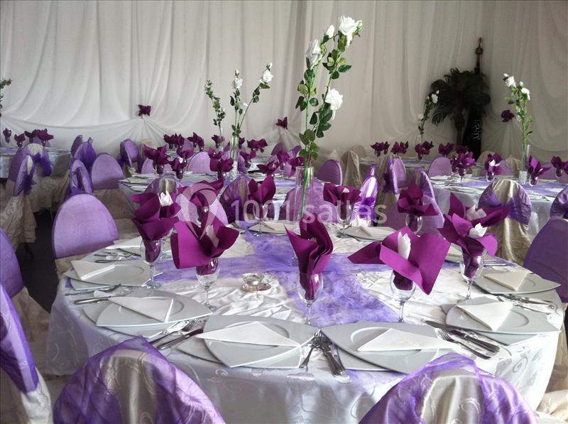 Salle décorée pour un événement avec des tables dressées, nappes blanches et violettes, serviettes pliées et fleurs blanches.