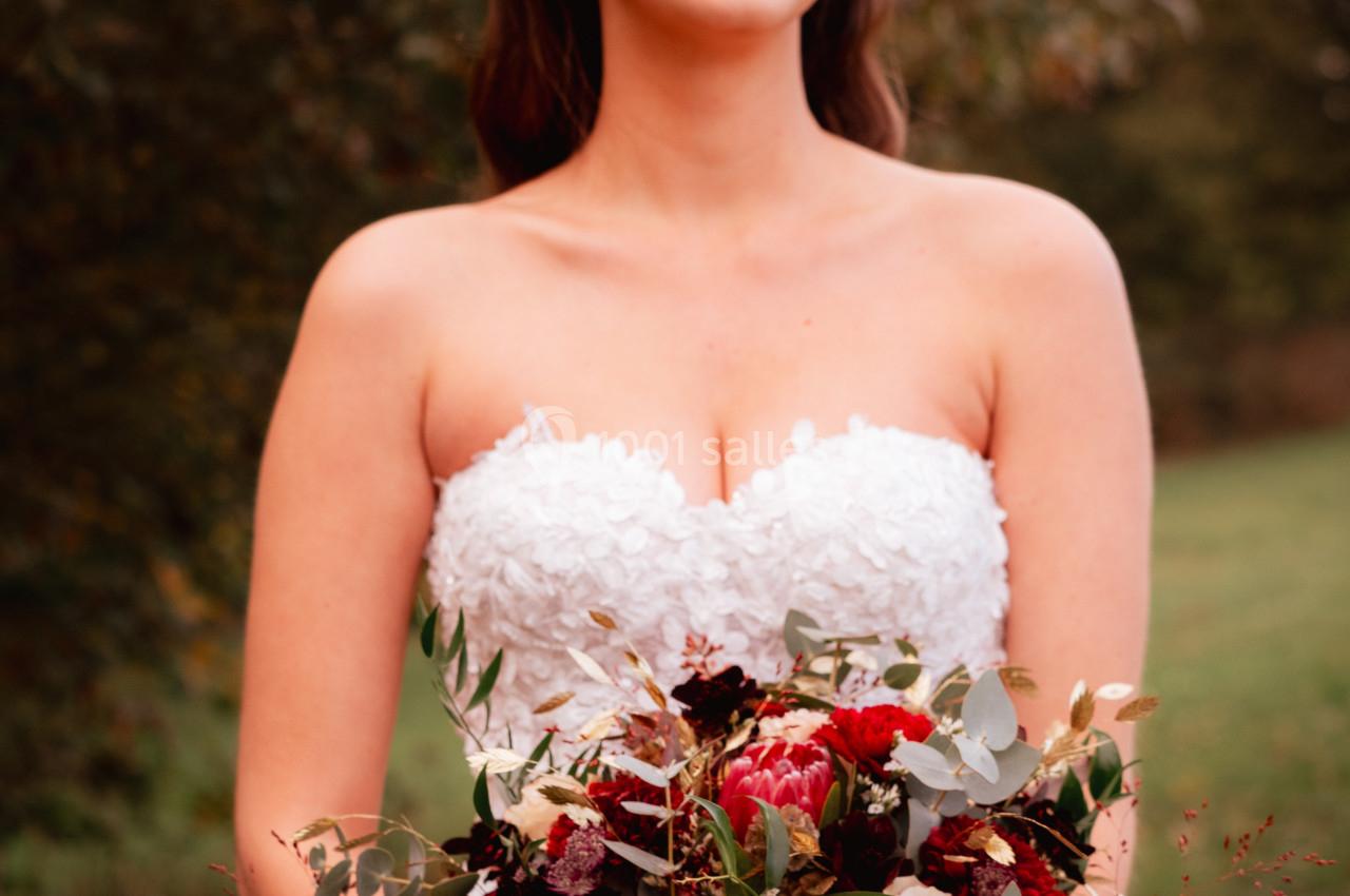 Une femme en robe de mariée tient un bouquet de fleurs rouges et blanches dans un cadre naturel.