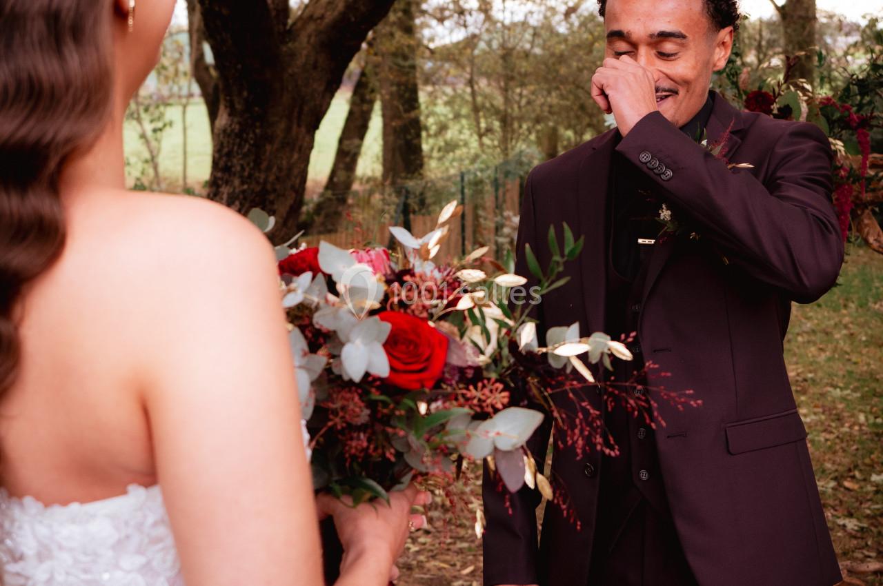 Un homme ému en costume sombre regarde une femme en robe blanche tenant un bouquet de fleurs rouges, dans un cadre boisé.