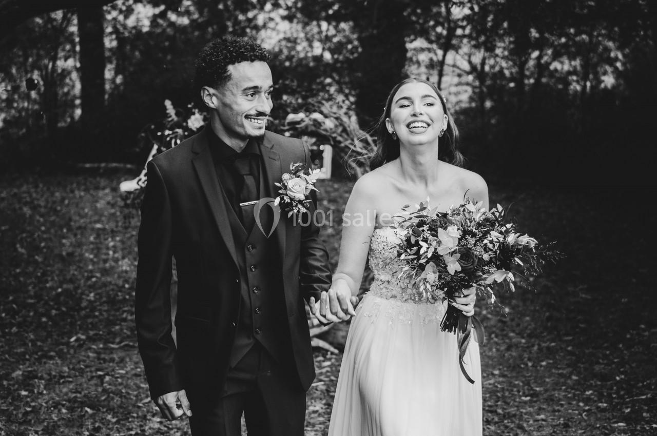 Un couple souriant marche main dans la main dans un cadre extérieur boisé, vêtus de tenues de mariage.