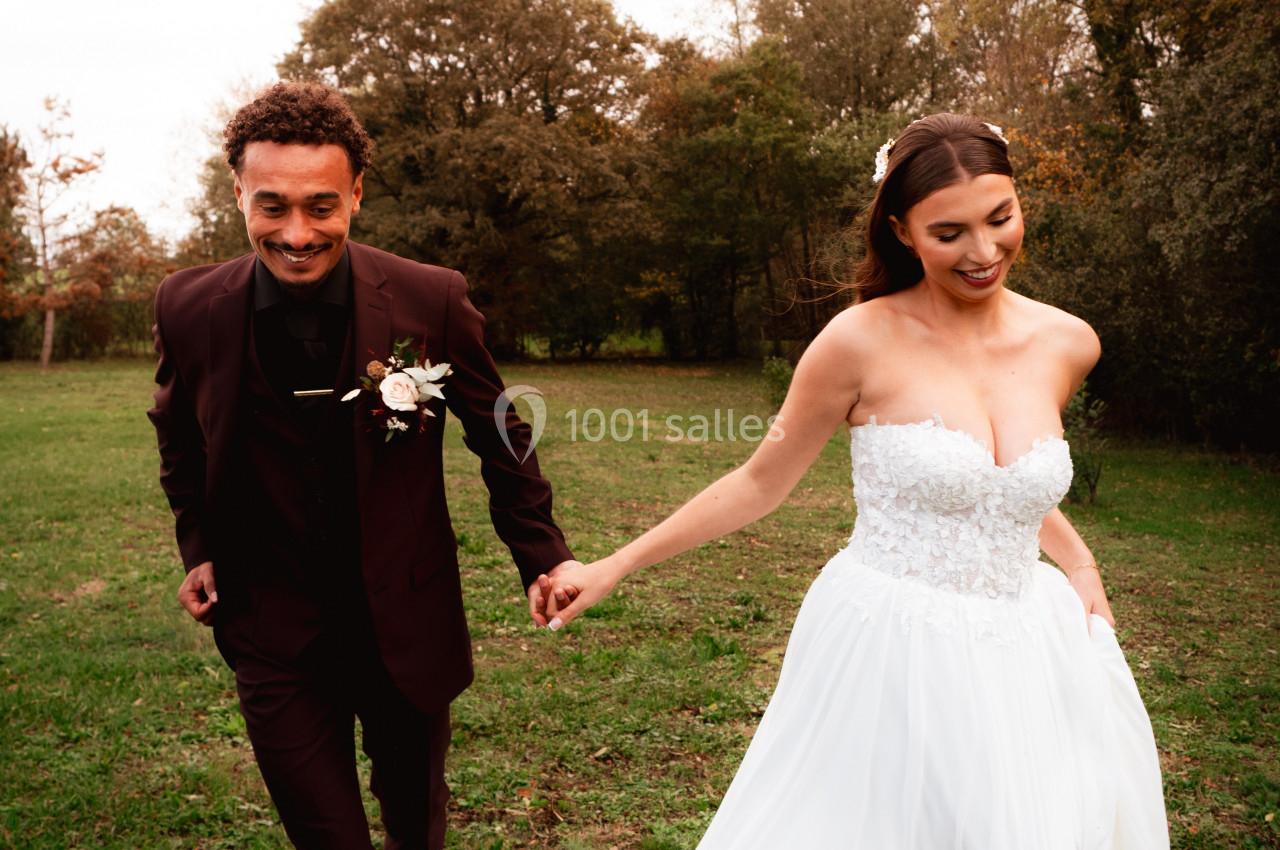Un couple en tenue de mariage marche main dans la main dans un champ verdoyant sous un ciel légèrement nuageux.