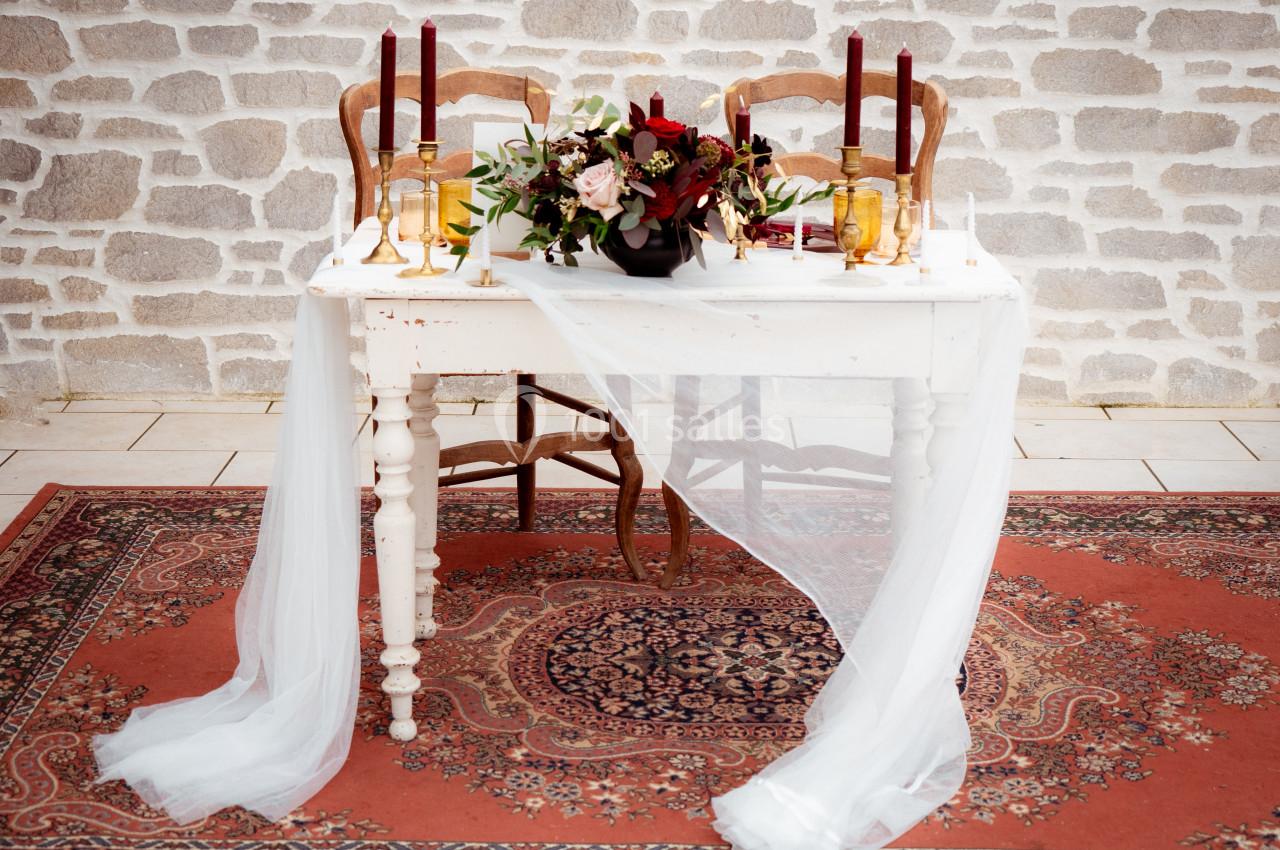 Table décorée avec des bougies rouges, un bouquet floral et un voile blanc, posée sur un tapis à motifs devant un mur en…
