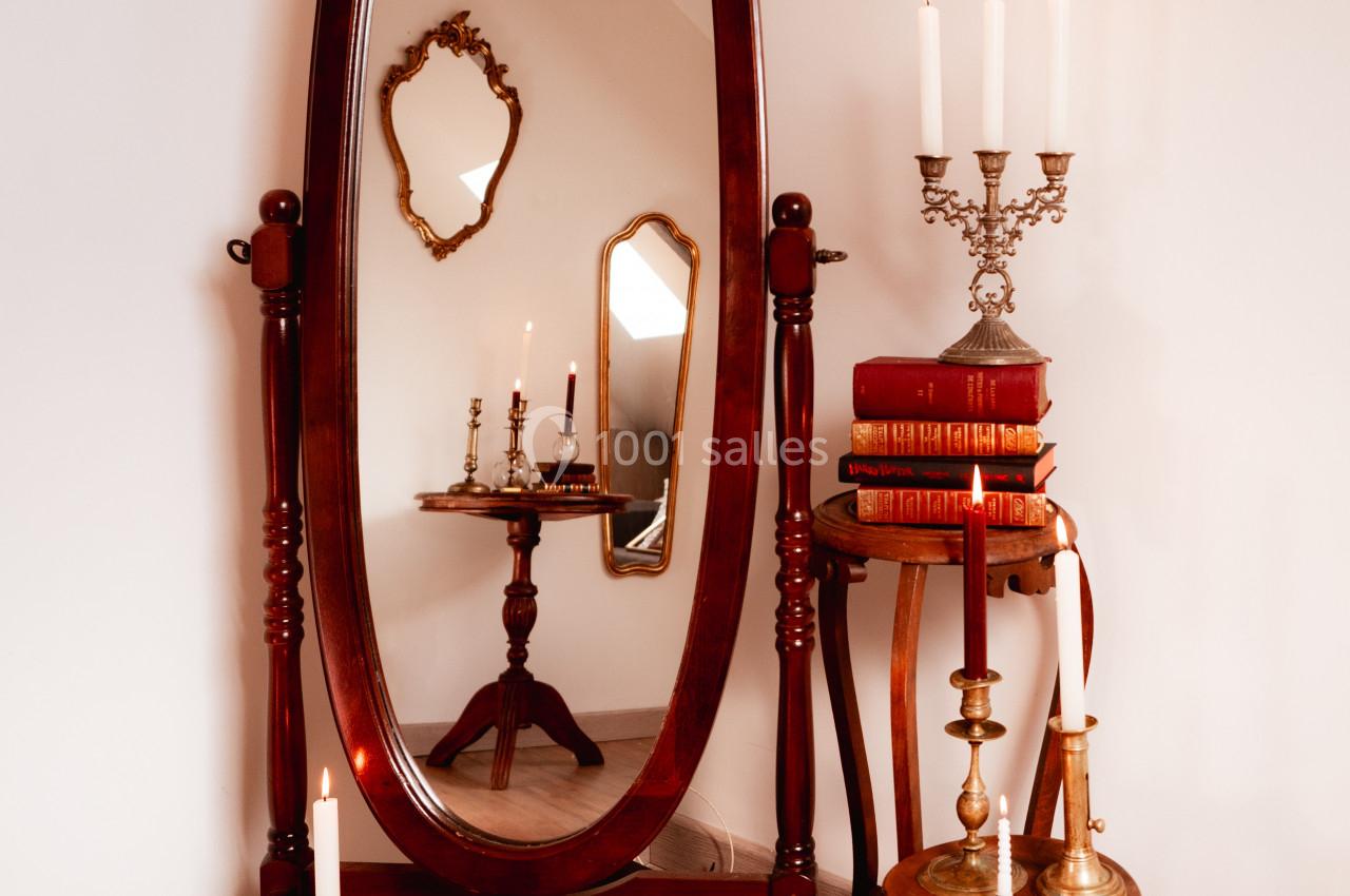 Grand miroir ovale sur pied entouré de bougies, livres anciens et meubles en bois dans un intérieur chaleureux.
