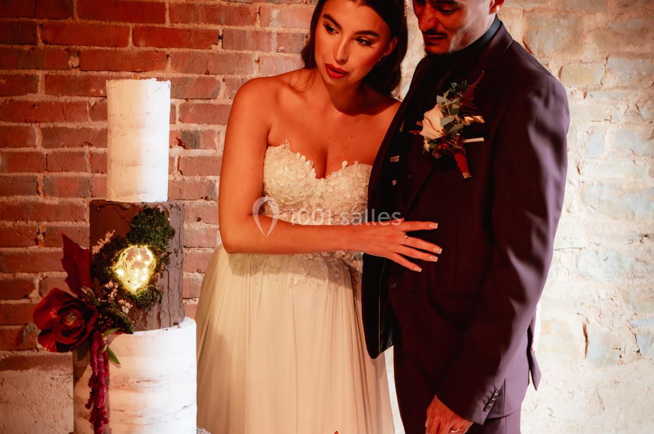 Un couple en tenue de mariage observe un gâteau à étages décoré de fleurs, dans un décor rustique en briques.