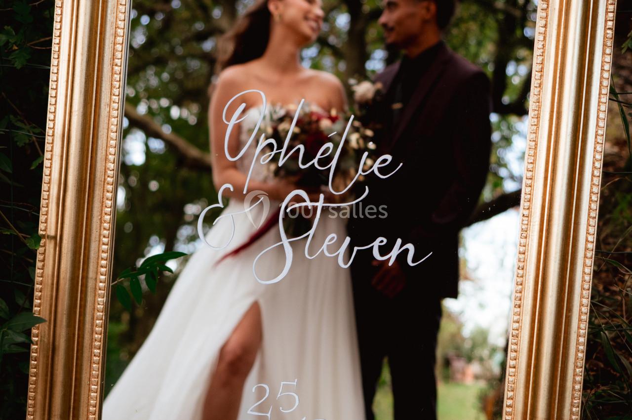Un couple souriant se reflète dans un miroir doré entouré de végétation, avec des inscriptions manuscrites sur la surface.