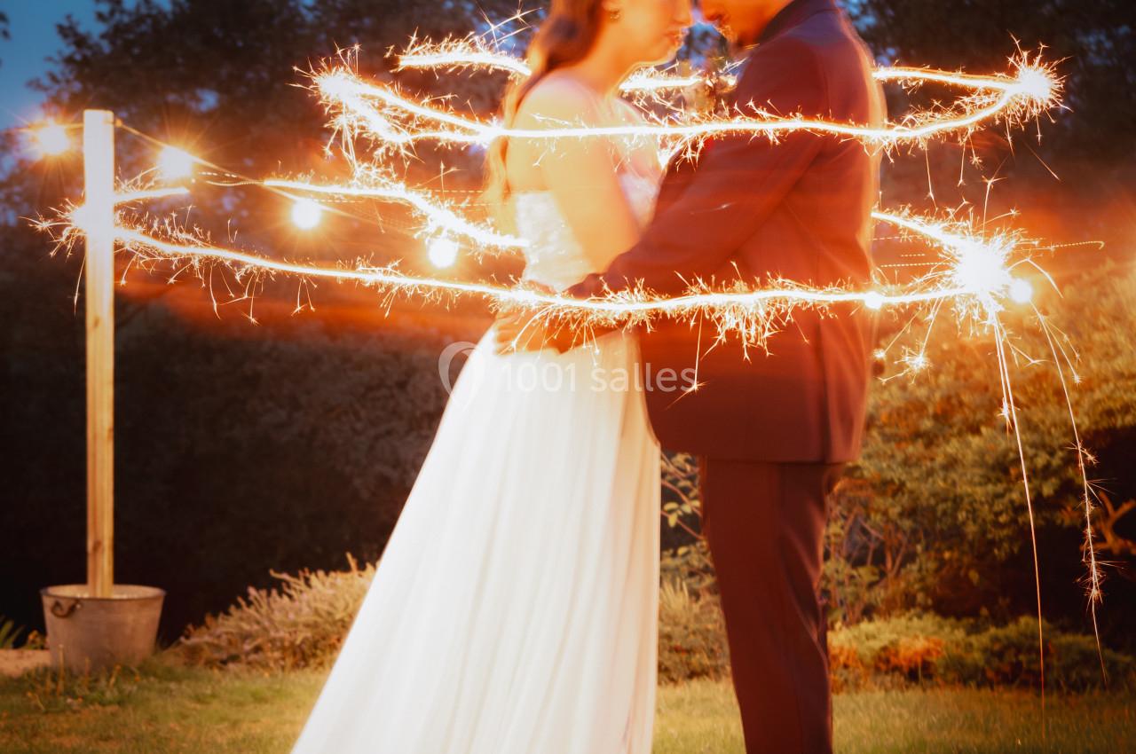 Un couple en tenue de mariage s'enlace dans un jardin, entouré d'étincelles lumineuses au crépuscule.