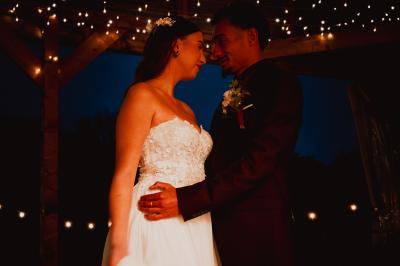 Un couple en tenue de mariage se regarde sous une pergola illuminée de guirlandes lumineuses.