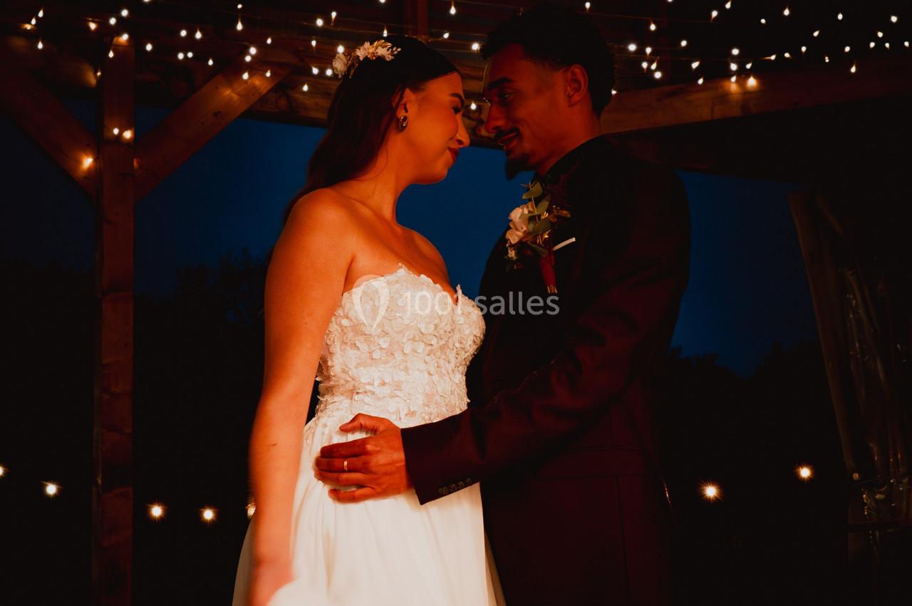 Un couple en tenue de mariage se regarde sous une pergola illuminée de guirlandes lumineuses.