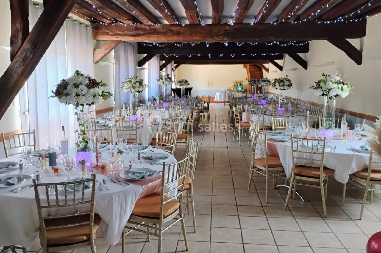 Salle de réception décorée avec des tables dressées, chaises dorées et arrangements floraux sous un plafond en poutres…