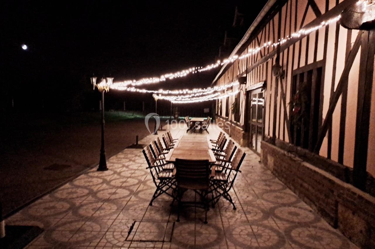 Grande table en bois entourée de chaises sur une terrasse éclairée par des guirlandes lumineuses, de nuit.