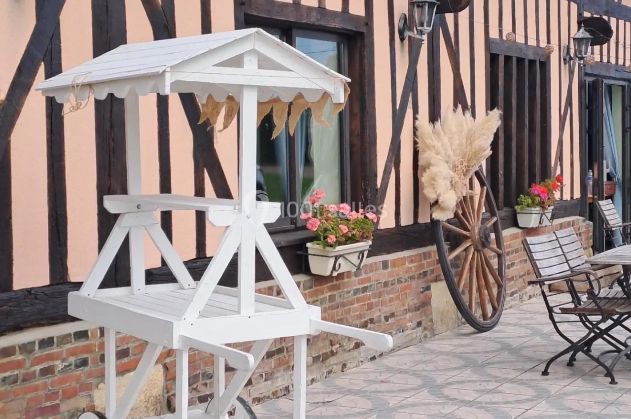 Charrette blanche en bois sur une terrasse pavée devant une maison à colombages avec décorations florales et rustiques.