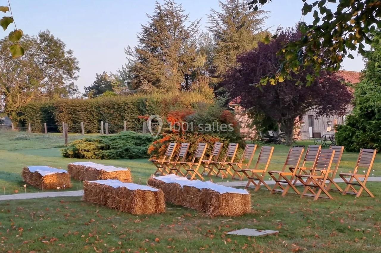 Chaises en bois et bottes de paille disposées en extérieur sur une pelouse, entourées d'arbres et de haies.