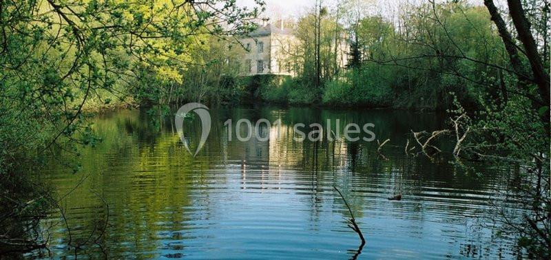 Vue d'un étang entouré de végétation avec un bâtiment blanc visible en arrière-plan à travers les arbres.