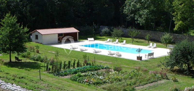 Vue d'une piscine extérieure entourée de transats, située dans un grand jardin avec potager et arbres.