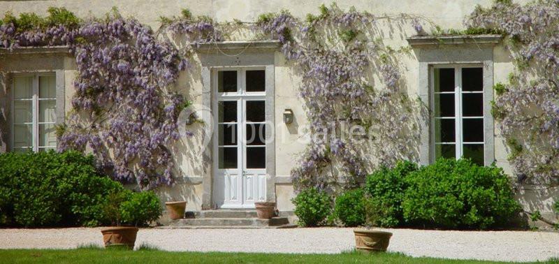 Façade d'une maison avec des murs recouverts de glycines en fleurs, entourée de buissons et de pots de fleurs.